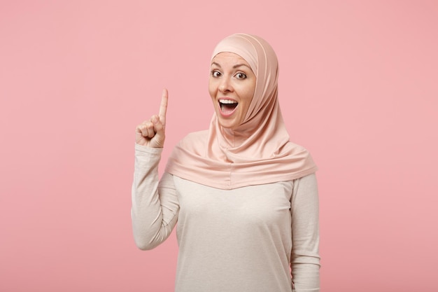 Eccitata giovane donna musulmana araba in abiti leggeri hijab in posa isolata su sfondo rosa. Concetto di stile di vita dell'Islam religioso della gente. Mock up spazio di copia. Alzando il dito indice con una nuova fantastica idea.