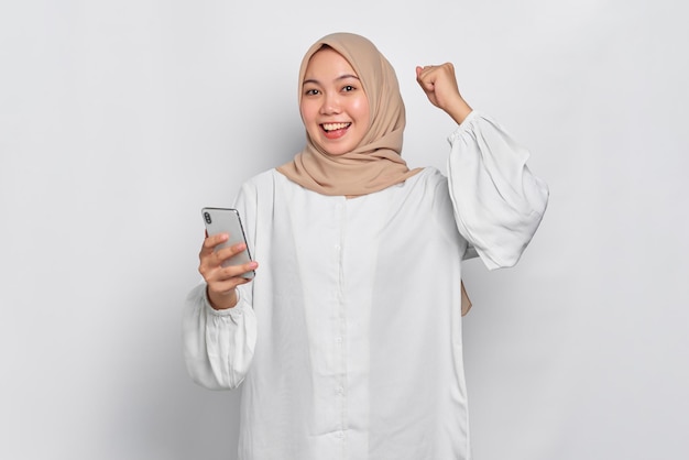 Eccitata giovane donna musulmana asiatica che usa un telefono cellulare e celebra il successo ottenendo buone notizie isolate su sfondo bianco