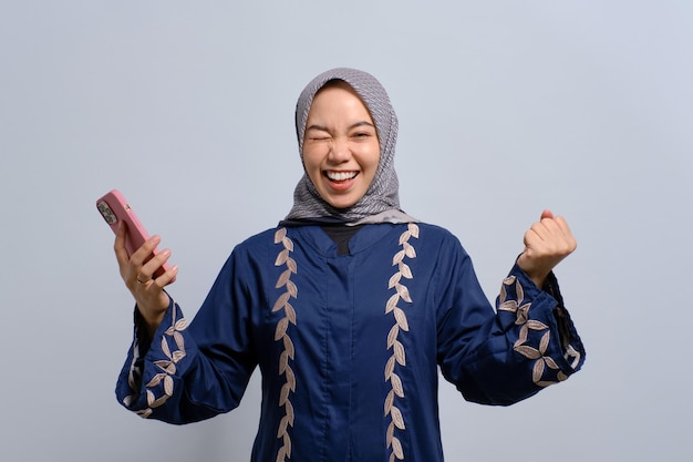 Eccitata giovane donna musulmana asiatica che utilizza il telefono cellulare e celebra il successo ottenendo buone notizie isolate su sfondo bianco