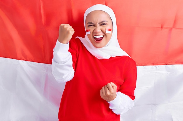Eccitata giovane donna musulmana asiatica in maglietta bianca rossa che celebra il giorno dell'indipendenza indonesiana il 17 agosto isolato su sfondo bandiera indonesiana