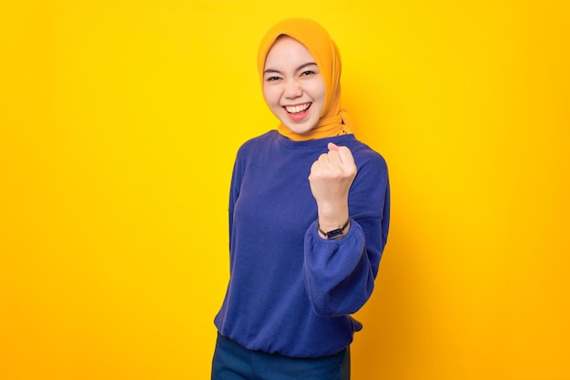 Eccitata giovane donna musulmana asiatica vestita con un maglione casual che celebra il successo con il pugno alzato isolato su sfondo giallo
