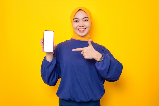 Eccitata giovane donna musulmana asiatica vestita con un maglione casual che mostra un telefono cellulare con schermo vuoto che consiglia un'app mobile isolata su sfondo giallo