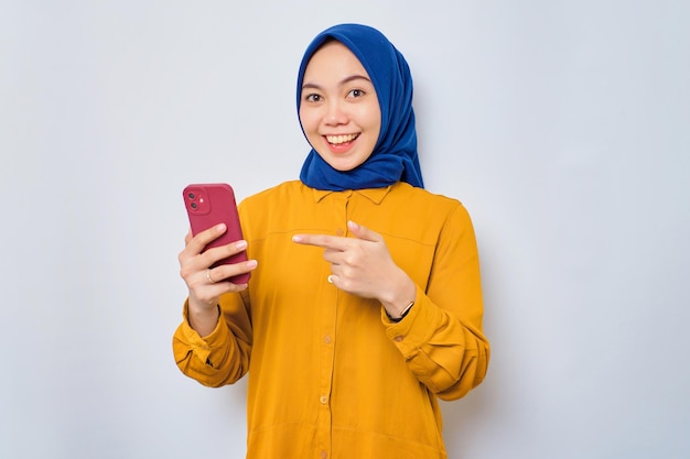 Eccitata giovane donna musulmana asiatica vestita con una camicia arancione che punta le dita verso il telefono cellulare che reagisce alla nuova app isolata su sfondo bianco