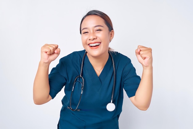 Eccitata giovane infermiera professionale asiatica femminile che lavora indossando un'uniforme blu in piedi fiduciosa mentre dice di sì e celebra la vittoria isolata su sfondo bianco Concetto di medicina sanitaria