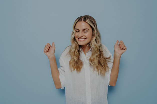 Eccitata giovane ragazza bionda piena di gioia esulta con entrambe le mani in aria, non può contenere la sua felicità esprimendo le sue emozioni con gesti mentre posa isolata sul muro blu. Concetto di eccitazione