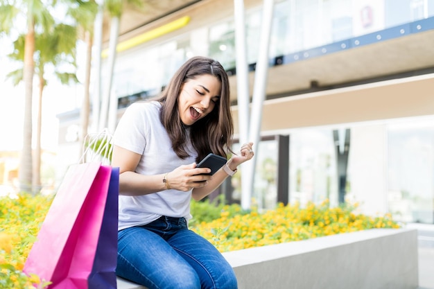 Eccitata metà donna adulta che utilizza lo smartphone con le borse della spesa fuori dal centro commerciale