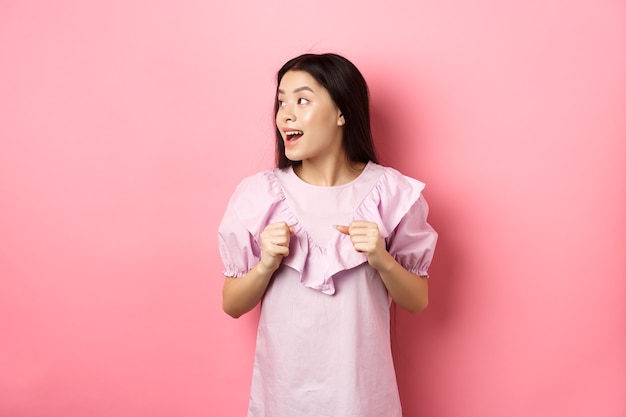 Eccitata ragazza asiatica guarda a sinistra con la faccia motivata, sorridendo felice, in piedi in abito su sfondo rosa.