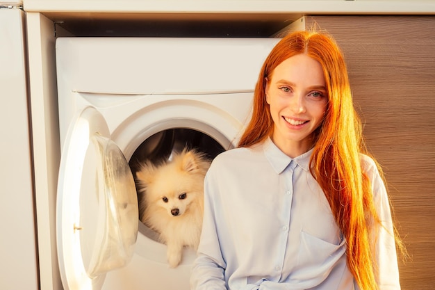 Eccitata ragazza dai capelli rossi divertente seduta accanto al suo adorabile spitz soffice all'interno della lavatrice a casa. giornata termale per il concetto di trattamento degli animali domestici.