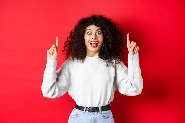 Eccitata ragazza moderna che mostra promo, puntando le dita verso l'alto e sorridendo stupita, raccontando grandi notizie, in piedi su sfondo rosso.