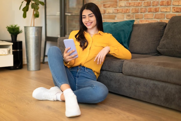 Eccitata signora adolescente spagnola che usa lo smartphone seduta sul pavimento vicino al divano nello spazio libero interno del soggiorno