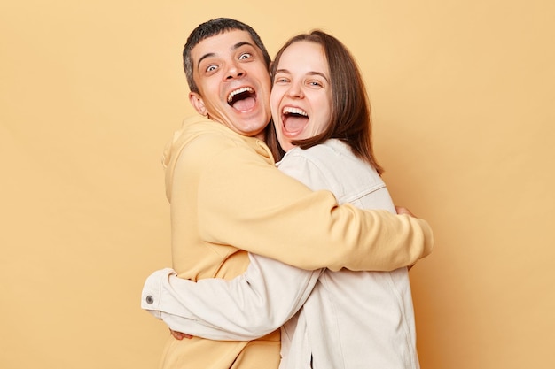 Eccitati amici felici donna e uomo che indossano abiti in stile casual in piedi isolati su sfondo beige che si abbracciano e ridono allegramente