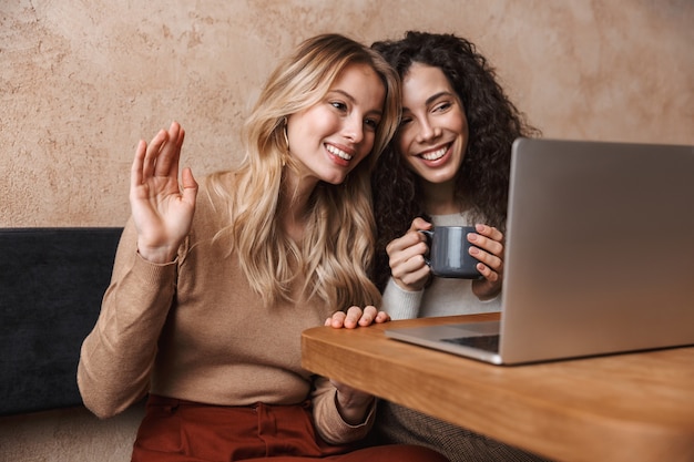 eccitati felici ragazze graziose amiche sedute al bar utilizzando il computer portatile che salutano parlando