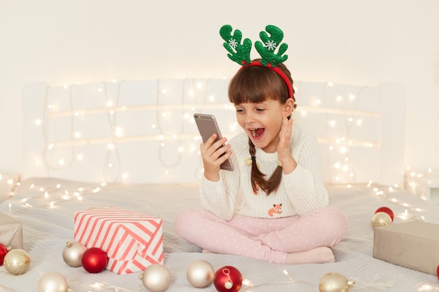 Eccitato affascinante ragazzina dai capelli scuri con le trecce che tiene il cellulare in mano, fare videochiamate o guardare video, urlare felicemente, avere grandi notizie sulla celebrazione delle vacanze invernali.