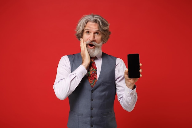 Eccitato anziano uomo barbuto con baffi dai capelli grigi in camicia classica cravatta in posa isolata su sfondo rosso. Concetto di stile di vita delle persone. Mock up spazio di copia. Tenere il telefono cellulare con lo schermo vuoto vuoto.