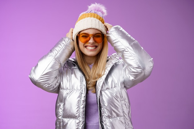 Eccitato audace impertinente giovane donna attraente divertirsi con gli amici viaggio invernale imparare lo snowboard sorridente sfacciato godendo la vacanza indossata cappello indossando occhiali da sole giacca argento caldo, sfondo viola.