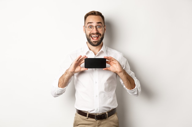 Eccitato bell'uomo che mostra lo schermo del telefono cellulare, guardando stupito, in piedi su sfondo bianco.