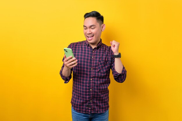 Eccitato giovane asiatico che usa lo smartphone e fa il gesto del vincitore isolato su sfondo giallo