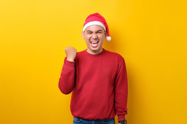 Eccitato giovane asiatico in cappello di Babbo Natale alzando i pugni per celebrare il successo su sfondo giallo studio celebrazione vacanze di Natale e concetto di Capodanno