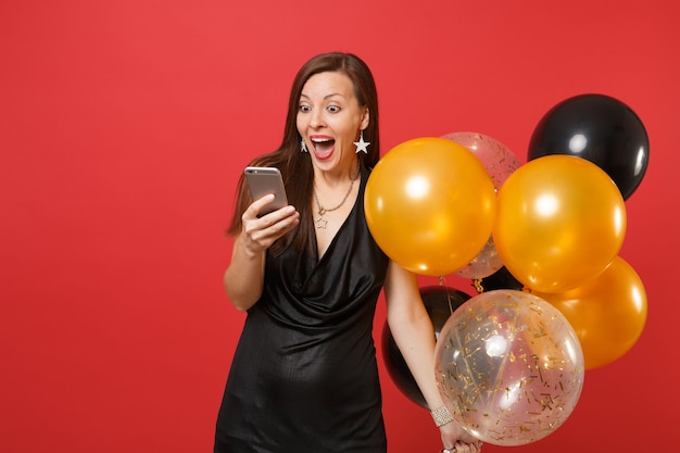 Eccitato giovane donna felice in abitino nero che celebra, tenendo i palloni ad aria e utilizzando il telefono cellulare isolato su sfondo rosso. Festa della donna, felice anno nuovo, concetto di festa di compleanno mockup.