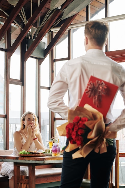 Eccitato giovane donna guardando il fidanzato con scatola di cioccolato e fiori che vengono al suo tavolo