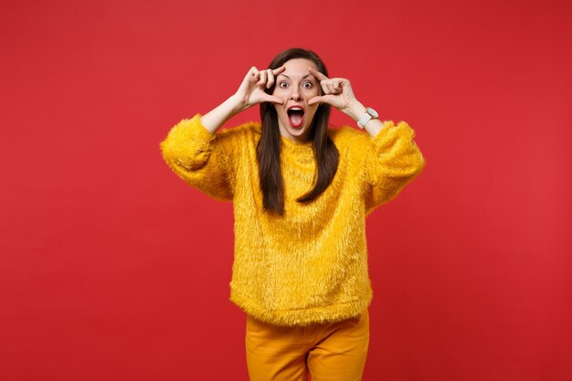 Eccitato giovane donna in maglione di pelliccia gialla che tiene la bocca spalancata, allungando le palpebre isolate su sfondo rosso brillante della parete in studio. Persone sincere emozioni, concetto di stile di vita. Mock up copia spazio.