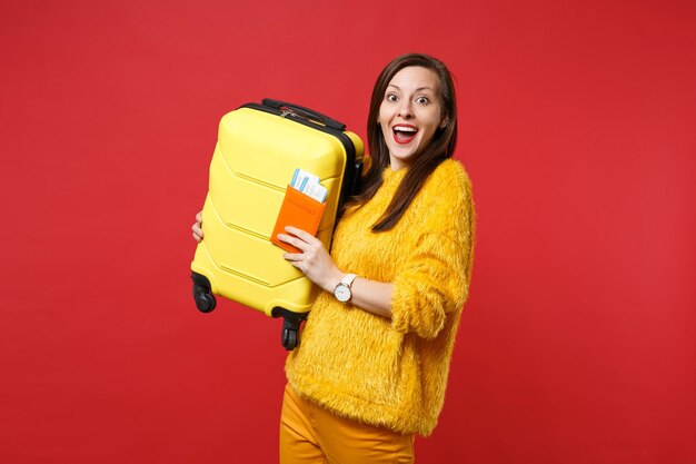 Eccitato giovane donna in maglione di pelliccia gialla che tiene la bocca spalancata tenere la valigia passaporto biglietto d'imbarco isolato su sfondo rosso. Concetto di stile di vita di emozioni sincere della gente. Mock up copia spazio.