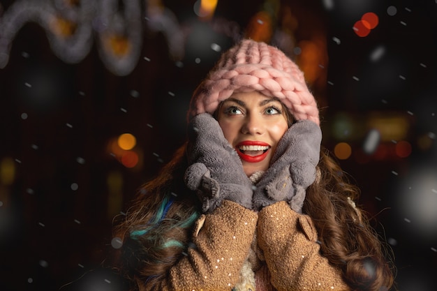 Eccitato giovane donna indossa cappello rosa, guanti e sciarpa a piedi in città la sera durante la nevicata