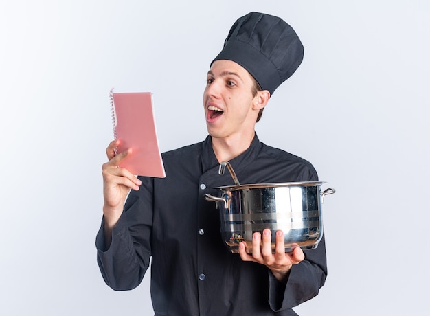 Eccitato giovane maschio biondo cuoco in uniforme da chef e cappello che tiene il blocco note di lettura della pentola isolato su parete bianca