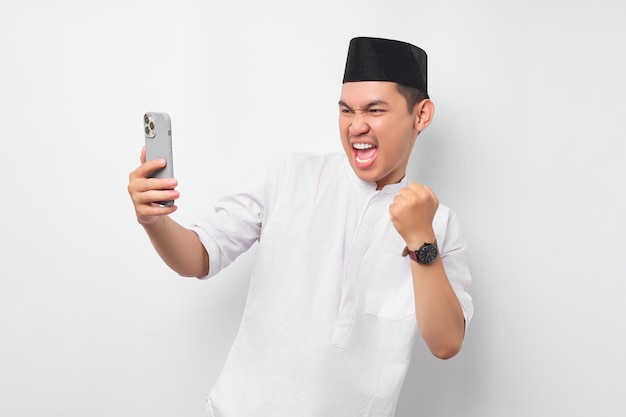 Eccitato giovane musulmano asiatico che utilizza il telefono cellulare e alza il pugno celebrando la buona fortuna isolato su sfondo bianco Persone religiose Islam stile di vita concetto celebrazione Ramadan e ied Mubarak