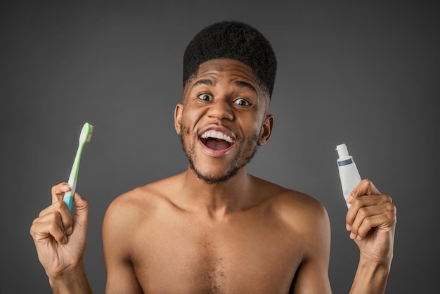 Eccitato giovane senza camicia che tiene spazzolino da denti e dentifricio isolati su sfondo grigio scuro