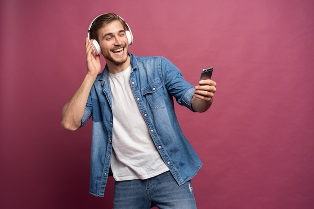 Eccitato giovane uomo che indossa una camicia di jeans in piedi isolato su sfondo rosa, ascoltando musica con gli auricolari e il telefono cellulare