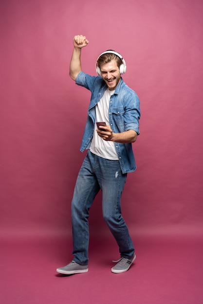 Eccitato giovane uomo che indossa una camicia di jeans in piedi isolato su sfondo rosa, ascoltando musica con gli auricolari e il telefono cellulare