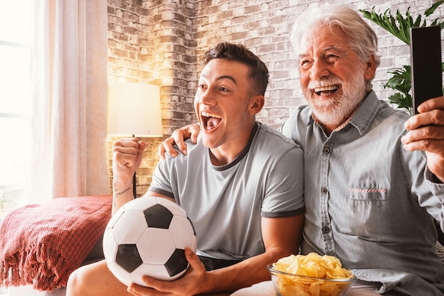 Eccitato tifoso di calcio del ragazzo che guarda una partita in televisione insieme a suo nonno che tiene in mano un pallone da calcio seduto su un comodo divano nel suo soggiorno