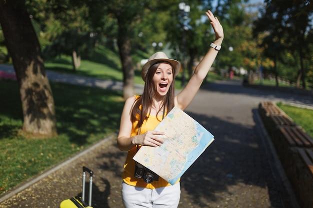 Eccitato turista donna viaggiatrice con valigia mappa della città agitando la mano per salutare, incontrare un amico, prendere un taxi in città all'aperto. Ragazza che viaggia all'estero per viaggiare nei fine settimana. Stile di vita del viaggio turistico.