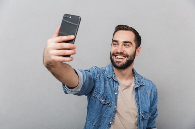 Eccitato uomo allegro che indossa una camicia isolata sopra il muro grigio, prendendo un selfie