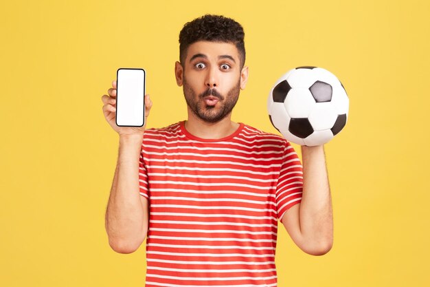 Eccitato uomo barbuto sorpreso in maglietta a righe con pallone da calcio e smartphone display bianco vuoto scioccato con una facile prenotazione del biglietto del campionato Colpo in studio al coperto isolato su sfondo giallo