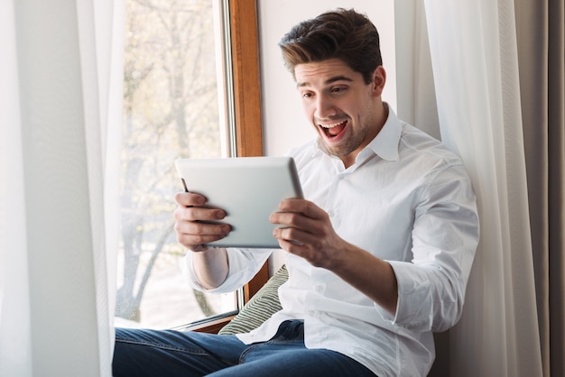 eccitato uomo caucasico che indossa una camicia bianca che gioca gioco sul computer tablet mentre era seduto alla finestra nel soggiorno