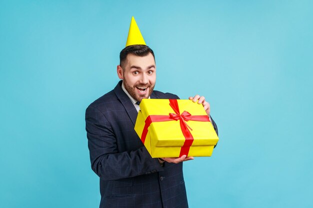 Eccitato uomo d'affari che indossa un abito in stile ufficiale e un cono di festa estremamente felice del regalo di festa, i sogni diventano realtà, guardando la fotocamera. Studio indoor girato isolato su sfondo blu.