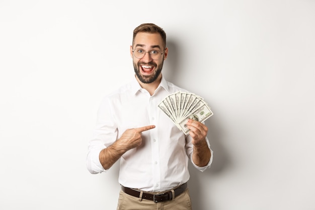 Eccitato uomo d'affari che punta al denaro, mostrando dollari e sorridente, in piedi su sfondo bianco.