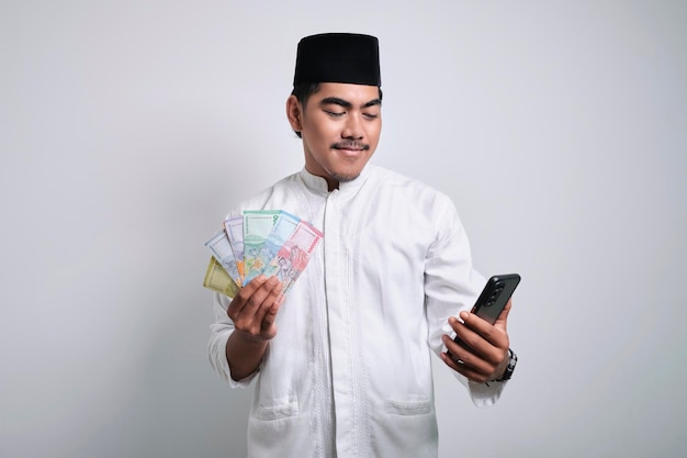 Eccitato uomo musulmano asiatico che indossa berretto e occhiali che tiene lo smartphone e mostra la mano piena di soldi