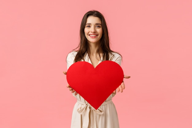 Ecco il mio amore per te. Affascinante tenera e seducente, romantica giovane donna che dà una grande carta del cuore, sorridendo felice, esprime il suo affetto e ammirazione, in piedi muro rosa