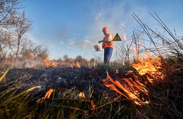 Ecologo vigile del fuoco che combatte un incendio in campo con cielo serale sullo sfondo Ambientalista maschio che tiene secchio e versa acqua sull'erba secca in fiamme vicino al segnale di avvertimento con punto esclamativo