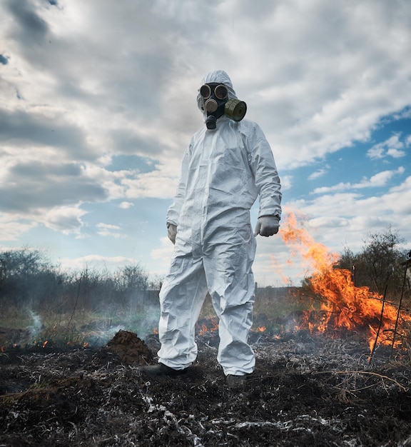 Ecologo vigile del fuoco che lavora in campo con incendio selvaggio Uomo in tuta protettiva contro le radiazioni e maschera a gas vicino a erba bruciante con fumo Concetto di disastro naturale