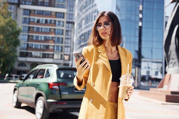 Edificio d'affari moderno sullo sfondo Giovane donna alla moda in cappotto color bordeaux durante il giorno con la sua auto