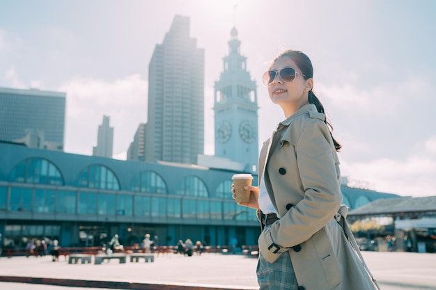 Edificio del traghetto di San Francisco con cielo blu soleggiato sullo sfondo. vista laterale del giovane lavoratore coreano asiatico sorridente dell'ufficio con una mano in tasca che cammina davanti a terra all'aperto. la donna beve il caffè