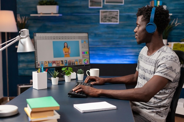 Editor di uomo afroamericano con le cuffie che fa ritocco fotografico sul computer utilizzando il software di post produzione della creatività. Fotografo che modifica la fotografia seduto alla scrivania in uno studio di ritocco domestico