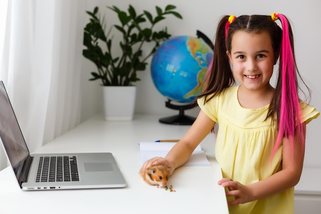 Educare a casa. La ragazza del bambino fa i compiti con il criceto dell'animale domestico. Criceto divertente dello zenzero che si siede sulla tavola in cui il bambino sta scrivendo. Di nuovo a scuola.