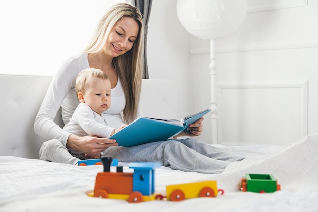 Educazione del bambino Madre felice con il suo bambino seduto sul letto e leggendo un libro