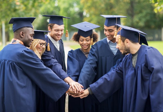 Educazione, laurea e concetto di persone - un gruppo di studenti internazionali felici in tavole di malta e abiti da scapolo che mettono le mani sopra