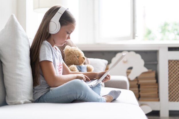 Educazione online dei bambini. Ragazza dolce con le cuffie che guarda il computer portatile di conferenza dell'insegnante di lezione video che si siede sul divano a casa.